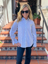 A woman wearing a light blue and white vertically striped button-down shirt with dark pants and sunglasses, standing on outdoor terracotta tile stairs with decorative tile accents.