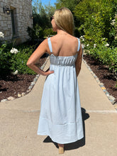 Woman in a blue and white striped dress standing on a sidewalk with greenery and flowers in the background.