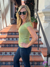 A woman wearing a light olive green short-sleeve t-shirt and dark blue jeans stands on outdoor terracotta steps with decorative tile borders, wearing black sunglasses.
