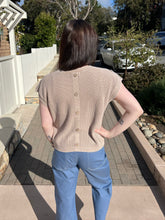 Woman wearing a khaki ribbed knit top standing on a sidewalk with trees and cars in the background