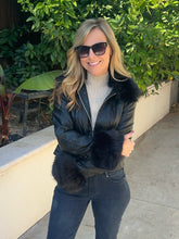 A woman wearing a black faux-fur-trimmed puffer jacket, dark sunglasses, a cream turtleneck, and dark jeans, standing outdoors on a concrete walkway with green foliage and a beige wall in the background.