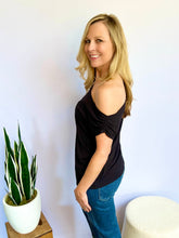 Woman wearing a black sleeveless top and blue jeans standing next to a potted plant against a white wall.