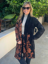 A woman wearing a long open-front cardigan with a black and rust-brown floral pattern, styled over a cream turtleneck and dark jeans, standing outdoors in front of a wall and green plants.