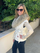 A woman wearing a cream quilted vest with a large gray faux-fur collar and a matching tie belt, paired with a beige ribbed knit sweater, dark pants, and sunglasses, standing outdoors in front of a beige wall and green foliage.