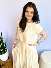 Woman wearing a yellow striped top standing indoors with a plant and box in the background.