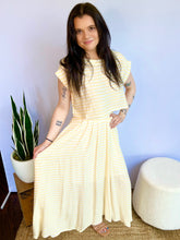 Woman wearing a yellow striped top standing indoors with a plant and box in the background.