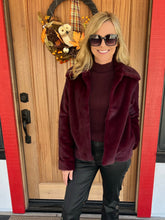 A woman wearing a plush burgundy faux fur jacket over a matching burgundy turtleneck sweater and black pants, standing in front of a wooden front door with a fall wreath.