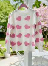 White sweater with pink heart patterns and a scalloped trim on a hanger outdoors.