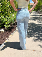 Person wearing light blue jeans standing on a sidewalk with greenery in the background