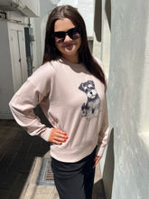 A woman wearing a light pink crewneck sweatshirt with a printed Schnauzer dog graphic on the front, paired with black pants and sunglasses, standing in a sunlit outdoor alleyway.