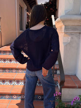 A woman viewed from behind wearing a dark navy blue open-knit sweater and blue jeans, standing on terracotta tiled steps in front of a white stucco building.