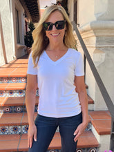 A woman wearing a white short-sleeve V-neck t-shirt and dark blue jeans, standing on outdoor tiled steps in front of a building.