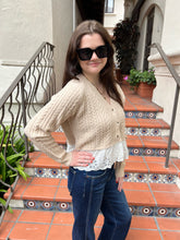A woman wearing a beige cable-knit cropped cardigan over a white lace top, paired with blue jeans and black sunglasses, standing on terracotta tiled outdoor stairs.