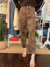 A person wearing brown cropped jeans with a dark brown abstract floral or marbled pattern, styled with a dark brown top and beige heeled sandals in a boutique setting.