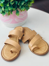 Pair of beige sandals on a white surface with a potted plant in the background.
