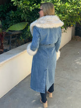 A woman wearing a long light blue denim coat with a large white faux-fur collar and matching white faux-fur trim at the cuffs, photographed outdoors on a concrete walkway with green plants in the background.