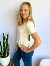 Woman wearing an ivory V-neck t-shirt and blue jeans standing next to a potted plant against a light blue wall.