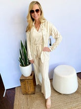 Woman wearing a striped shirt and blue jeans standing next to a potted plant against a light blue wall.