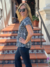 A woman wearing a black and white floral patterned short-sleeve blouse and dark jeans, standing on outdoor terracotta tile steps with decorative blue mosaic tiles.