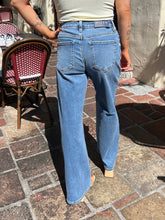 A person shown from the back wearing light blue wide-leg denim jeans, standing on a brick patio with a woven chair and wall in the background.
