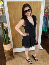 Woman in a black dress posing in front of a wall with a cactus plant and clothing rack in the background.