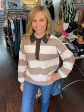 A smiling blonde woman wearing a brown and cream striped knit polo sweater with blue jeans, standing inside a clothing boutique.