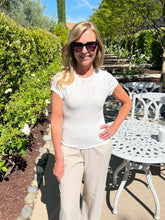 Woman in a white top and beige pants standing outdoors with greenery and a white table in the background.