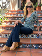 A woman wearing a blue and white patterned V-neck top, dark blue jeans, and tan wedge sandals sits on terracotta tiled steps with decorative blue and white tiles.
