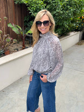 A woman wearing a sheer, long-sleeve high-neck blouse with a gray and white abstract pattern, paired with blue jeans and black sunglasses, standing outdoors on a concrete walkway with green plants and a wooden fence in the background.