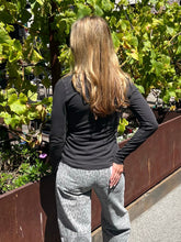 A woman viewed from behind wearing a black long-sleeve top and gray leopard-print pants, standing outdoors in front of green plants and a brown planter box.