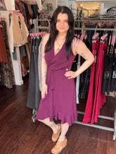 A woman modeling a sleeveless wrap-style midi dress in a rich plum purple color, standing inside a clothing boutique with racks of garments in the background.