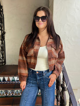 A woman wearing a brown and beige plaid cropped flannel shirt jacket over a cream tank top and blue jeans, standing in front of a textured white stucco wall.
