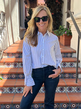 A woman wearing a light blue and white vertically striped button-down shirt with dark jeans and sunglasses, posing on outdoor terracotta tile steps.