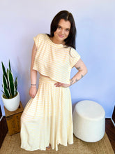 Woman wearing a yellow striped top standing indoors with a plant and box in the background.