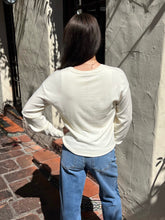 A person viewed from behind wearing a cream-colored long-sleeve crewneck top and blue jeans, standing outdoors against a textured stucco wall.