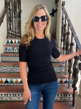 A woman with blonde hair and black sunglasses wearing a black short-sleeve crewneck top and blue jeans, standing on a tiled staircase.