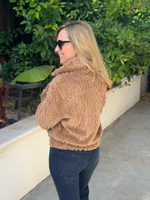 A woman with blonde hair and sunglasses, wearing a chunky tan cable-knit cowl-neck sweater and dark blue jeans, standing outdoors in front of a white wall and green foliage.