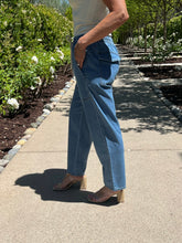 Person wearing blue jeans and a white top standing on a sidewalk with greenery in the background