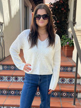 A woman wearing a cream cable-knit long-sleeve sweater and blue jeans stands on tiled outdoor stairs.