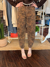 A person wearing brown acid-wash cropped jeans with a mottled distressed pattern, paired with beige block-heel sandals, standing in a clothing boutique.