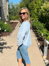 Woman in a light blue striped shirt and denim shorts standing on a sidewalk with greenery in the background.