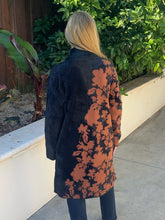 A woman viewed from behind wearing a long black coat with a large rust-orange floral print running down the back, standing outdoors on a concrete walkway with green plants and a white wall behind her.