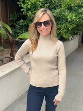 A woman wearing a beige ribbed turtleneck sweater and dark blue jeans, standing outdoors in front of green plants and a concrete wall.