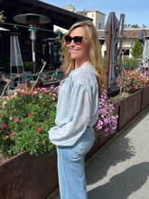 Woman in light blue top and jeans standing in a garden with flowers and outdoor furniture.