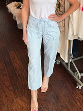 Person wearing light blue pants and a white top in a room with wooden floor and clothing rack.
