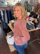 A smiling blonde woman wearing a pink and white vertically striped ribbed knit sweater and blue jeans, standing inside a clothing boutique.