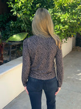 A woman viewed from behind wearing a long-sleeve crewneck top with a dark brown and black abstract leaf-like pattern, paired with dark blue jeans, standing outdoors on a concrete path with green foliage in the background.