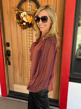 A woman wearing a sheer long-sleeve blouse with a rust red and orange diagonal dotted pattern, paired with black pants, standing in front of a wooden front door with a fall wreath.