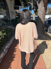Person wearing a natural sweater walking on a sidewalk with trees and parked cars in the background.