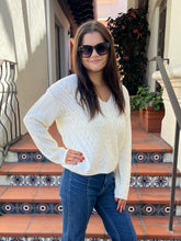 A woman wearing a cream cable-knit V-neck sweater and blue jeans stands on tiled outdoor stairs.